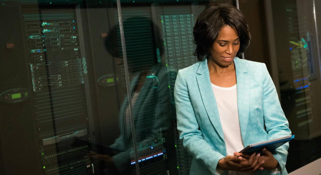 A woman in a turquoise blazer looks at a tablet while standing in front of a computer server.